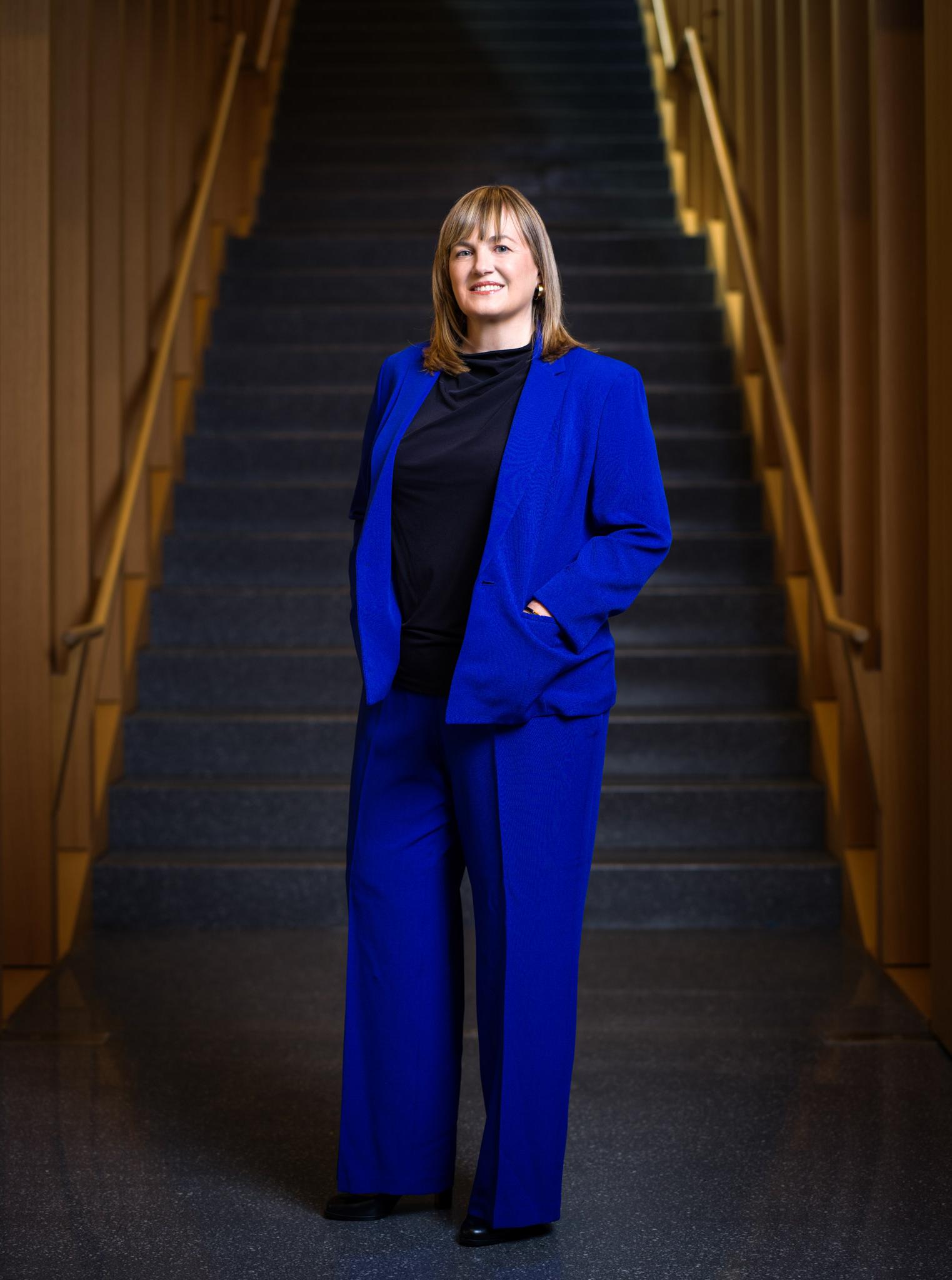 劳拉·罗森伯里总统站在米尔斯坦大楼的楼梯前. She is wearing a blue suit